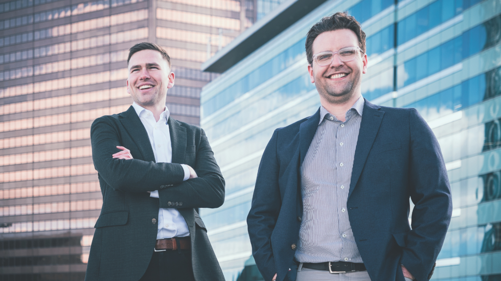 The image shows two individuals standing in front of modern office buildings. Both are dressed in business attire, with one wearing a dark suit and the other wearing a dark blazer with a light-colored shirt. The background consists of tall glass buildings reflecting the urban environment.